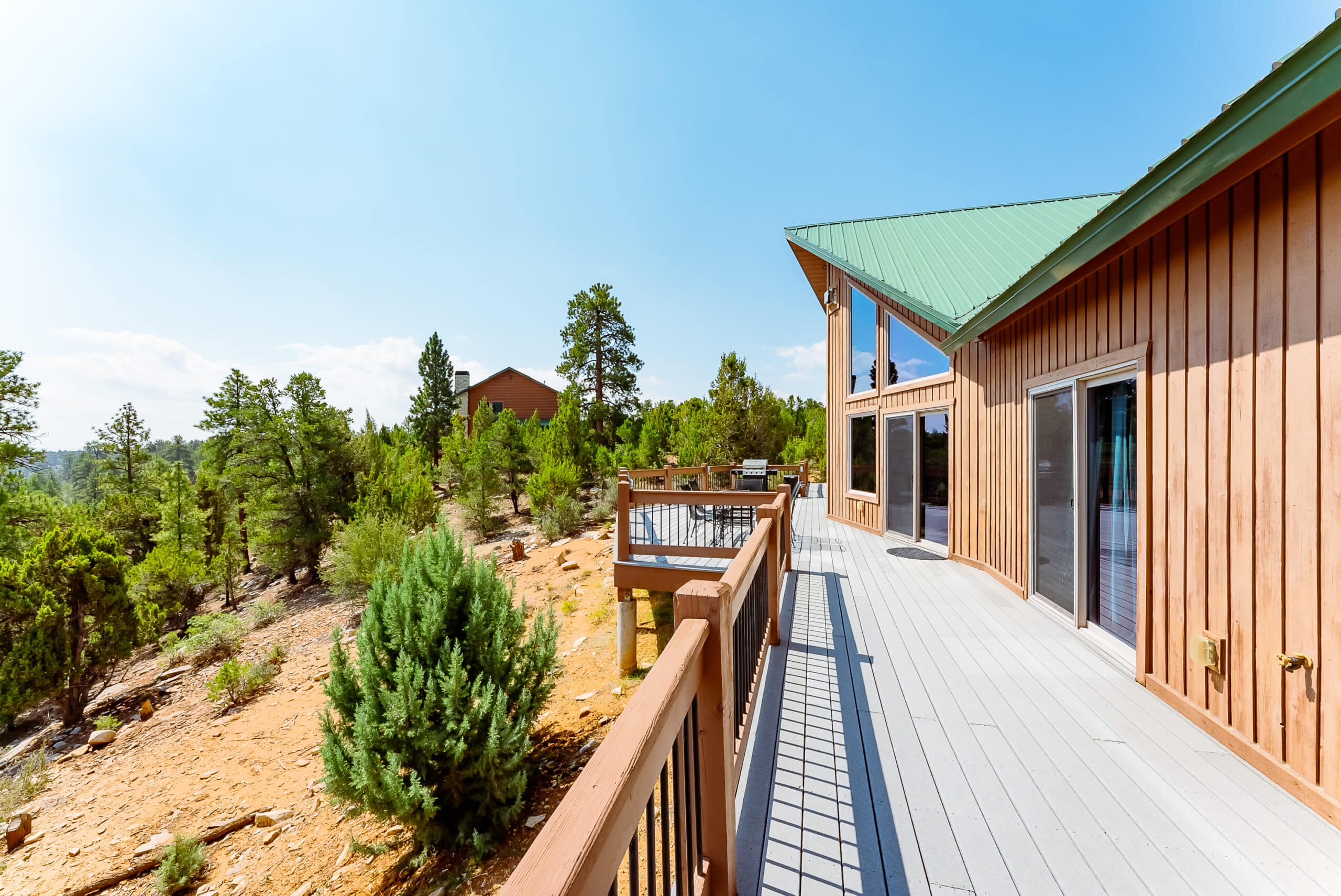 A wooden deck leads to a modern cabin surrounded by lush greenery and clear blue skies.